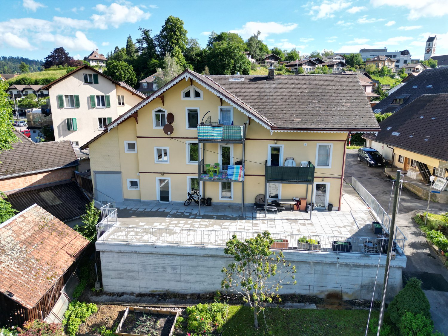 Two-story yellow house with balcony, surrounded by garden and other houses, clear sky above.