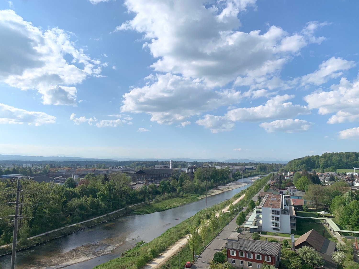 The image shows a scenic view of a city with a river running through it. The cityscape includes a mix of residential and commercial buildings, surrounded by lush greenery and a cloudy blue sky. The river appears to be a prominent feature, with a walking/c