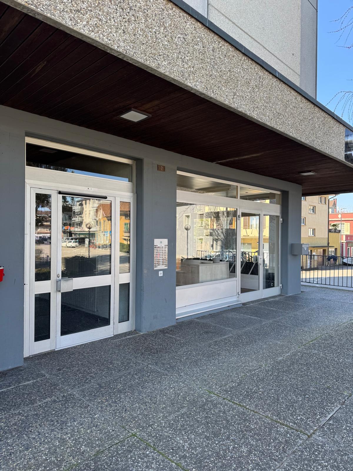 Exterior of a building with glass doors and windows, gray walls, and a paved entrance.