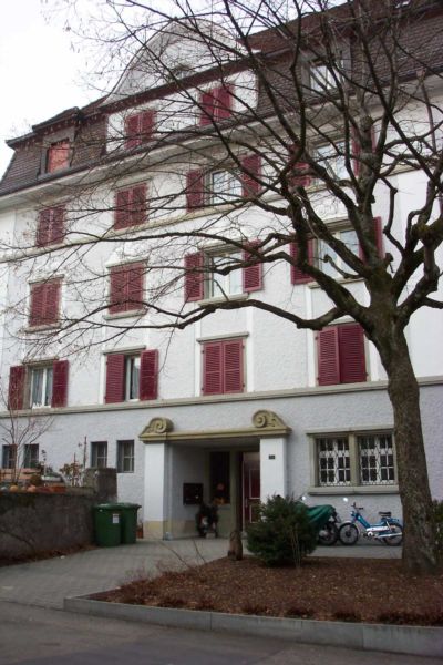 3-story white building with red shutters, a tree in the foreground, and a police car parked in front of the building.
