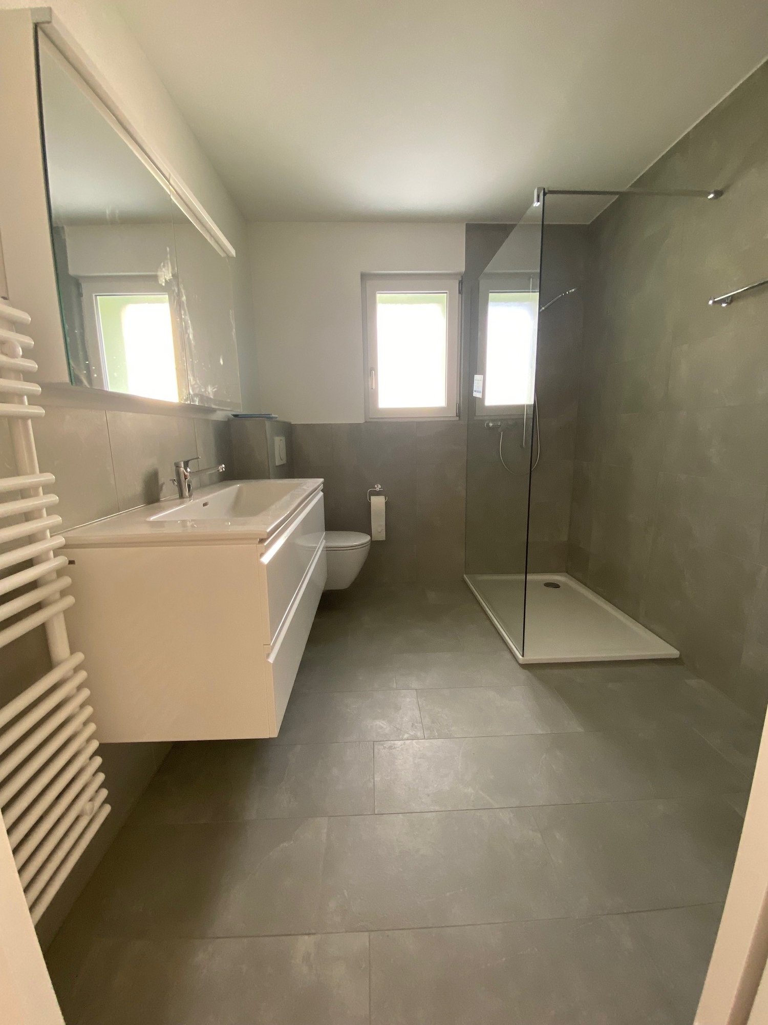 A modern bathroom with white fixtures, gray tiled floors, a glass walk-in shower with a showerhead, a white vanity with a sink and faucet, a toilet, a mirror above the vanity, and a towel rack