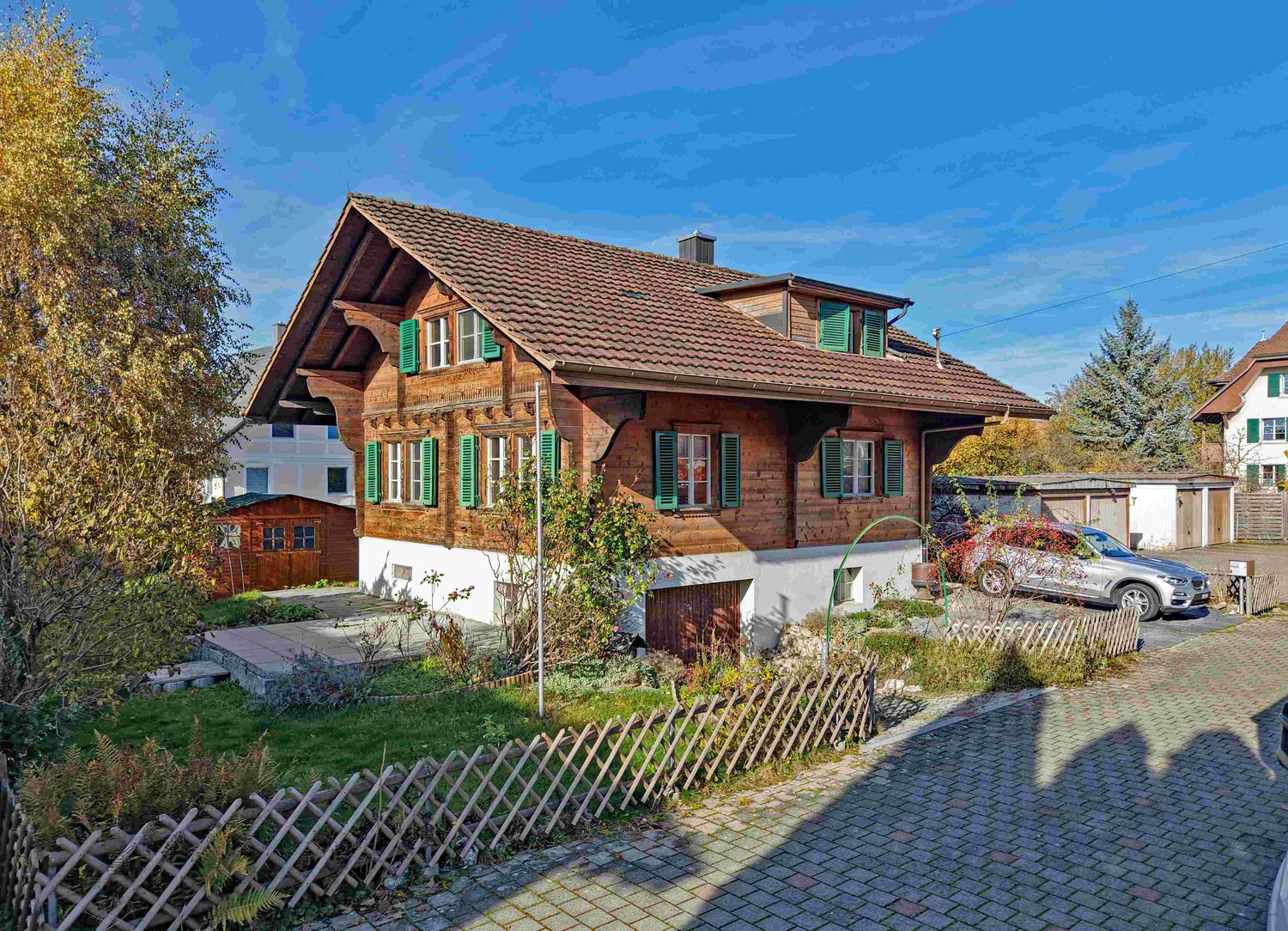 Detached house, brown wooden facade, green shutters, brown roof, small side building, driveway, car parked in front, brick sidewalk