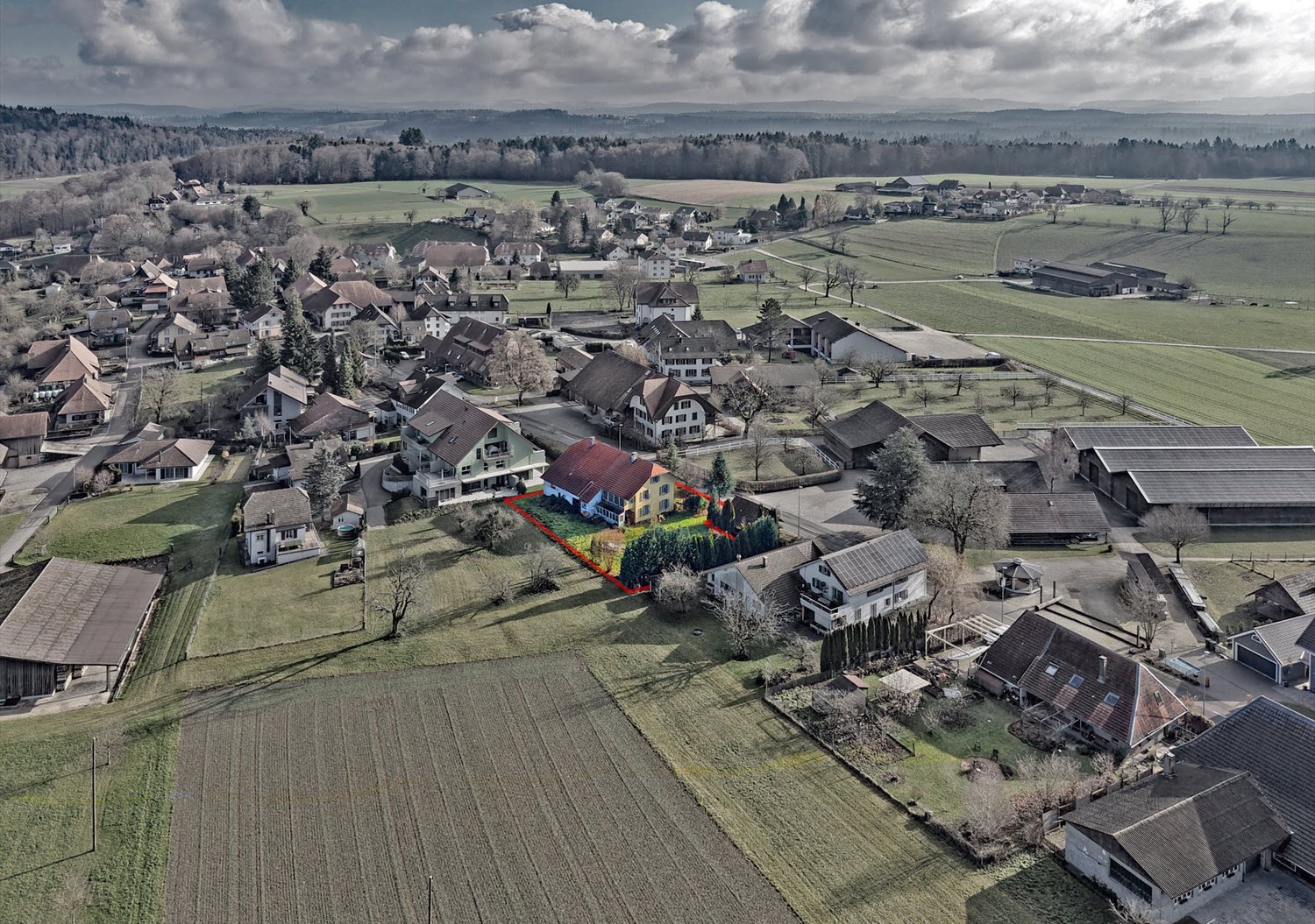 Rural area with scattered houses, central house highlighted, flat agricultural fields, sparse trees, wide view of surrounding countryside, some houses have solar panels
