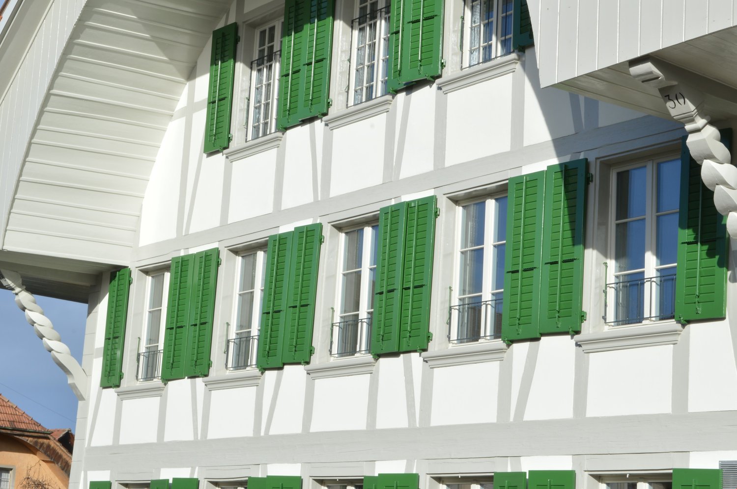 A two-story white building with green shutters on the windows. The building has a traditional architectural style with a pitched roof and decorative elements. There are several windows with green shutters on the facade.