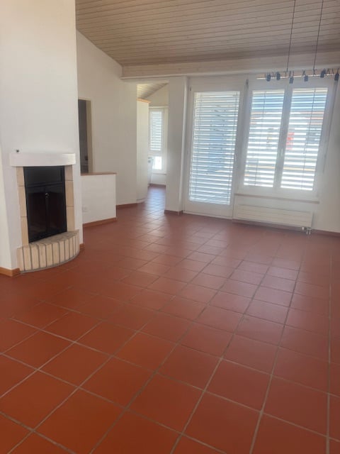 Spacious living room with terracotta tile flooring, white walls, and a fireplace. Large windows provide natural light and views. The room has an open layout with a high ceiling.