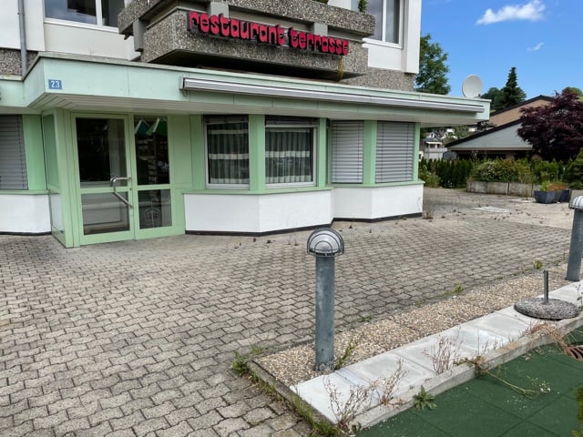 An establishment building with brick floors, a green exterior with white shutters, a glass entrance door, a trash basket, a round lamp on the side, and trees and plants on the side.