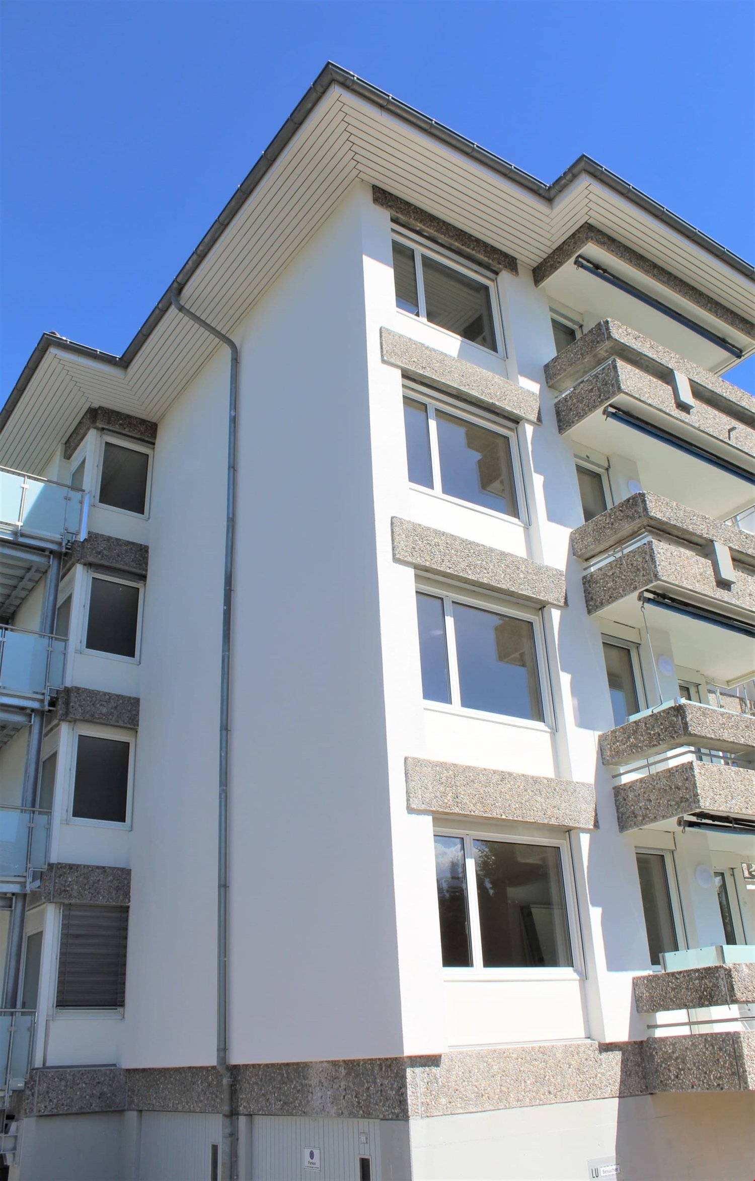 Multi-story residential building, white exterior, balconies on upper floors, large windows, modern architecture, grey stone accents, flat roof with ridge tiles