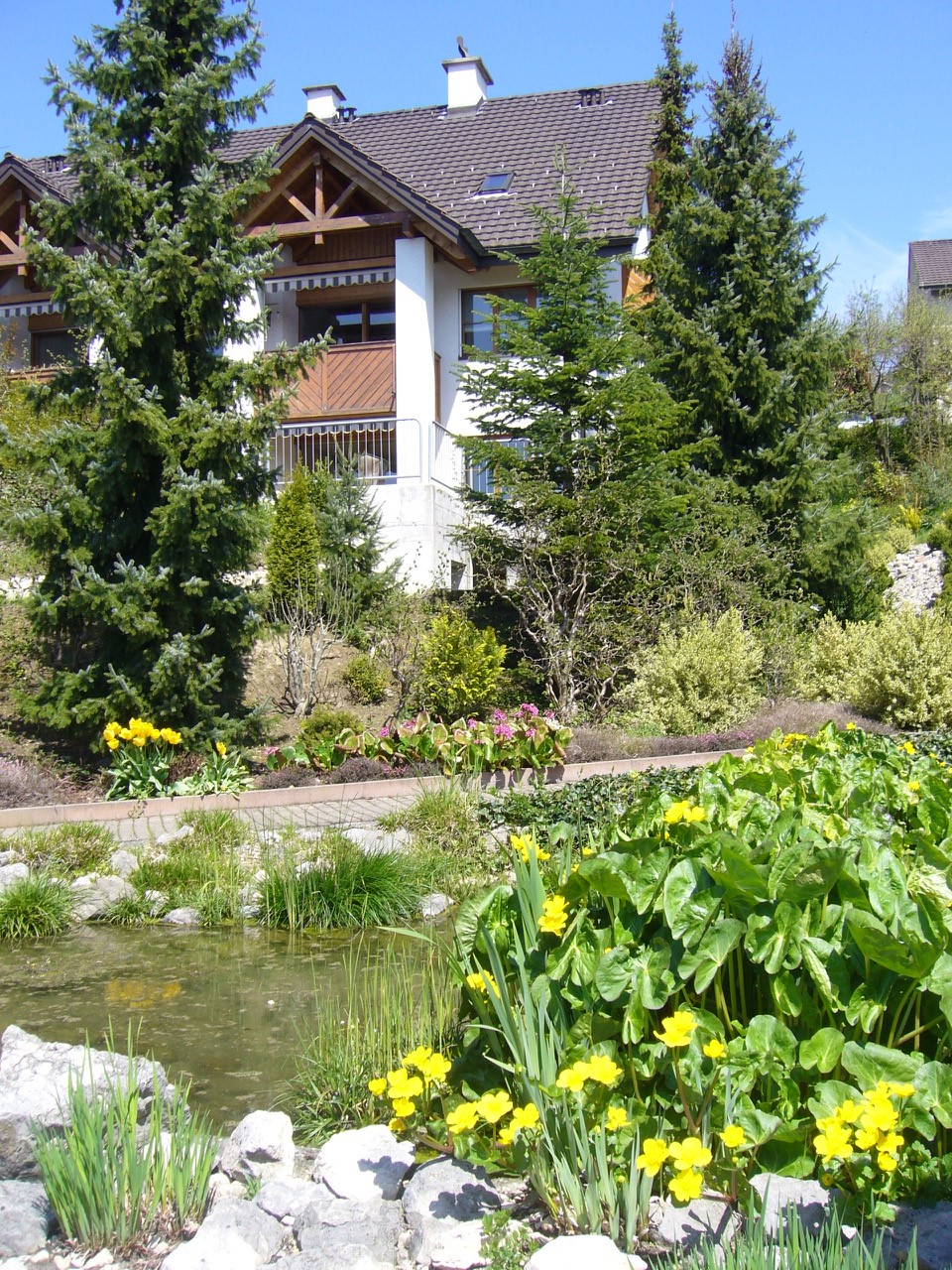 Exterior of two-story house with brown roof, white walls, wooden beams, large glass windows, balcony, garden with flowers, shrubs, trees, small pond, rocks