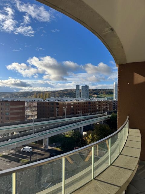 an outdoor balcony/terrace with a glass railing and a concrete surface, a view of an elevated highway with moving vehicles and distant city buildings