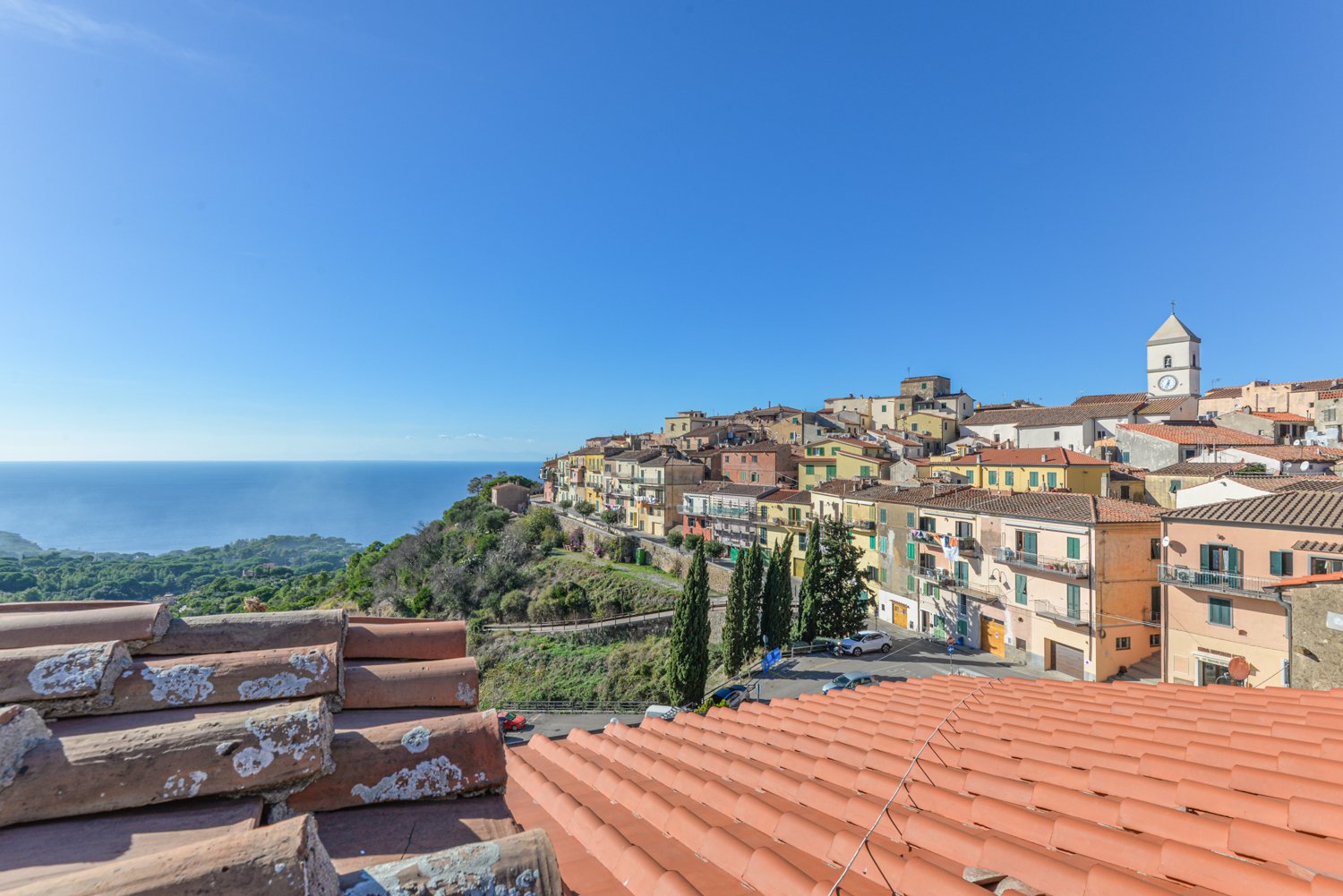 Picturesque coastal town with colorful buildings, red tile roofs, and a church steeple overlooking the sea. The town is situated on a hillside with lush greenery surrounding it.