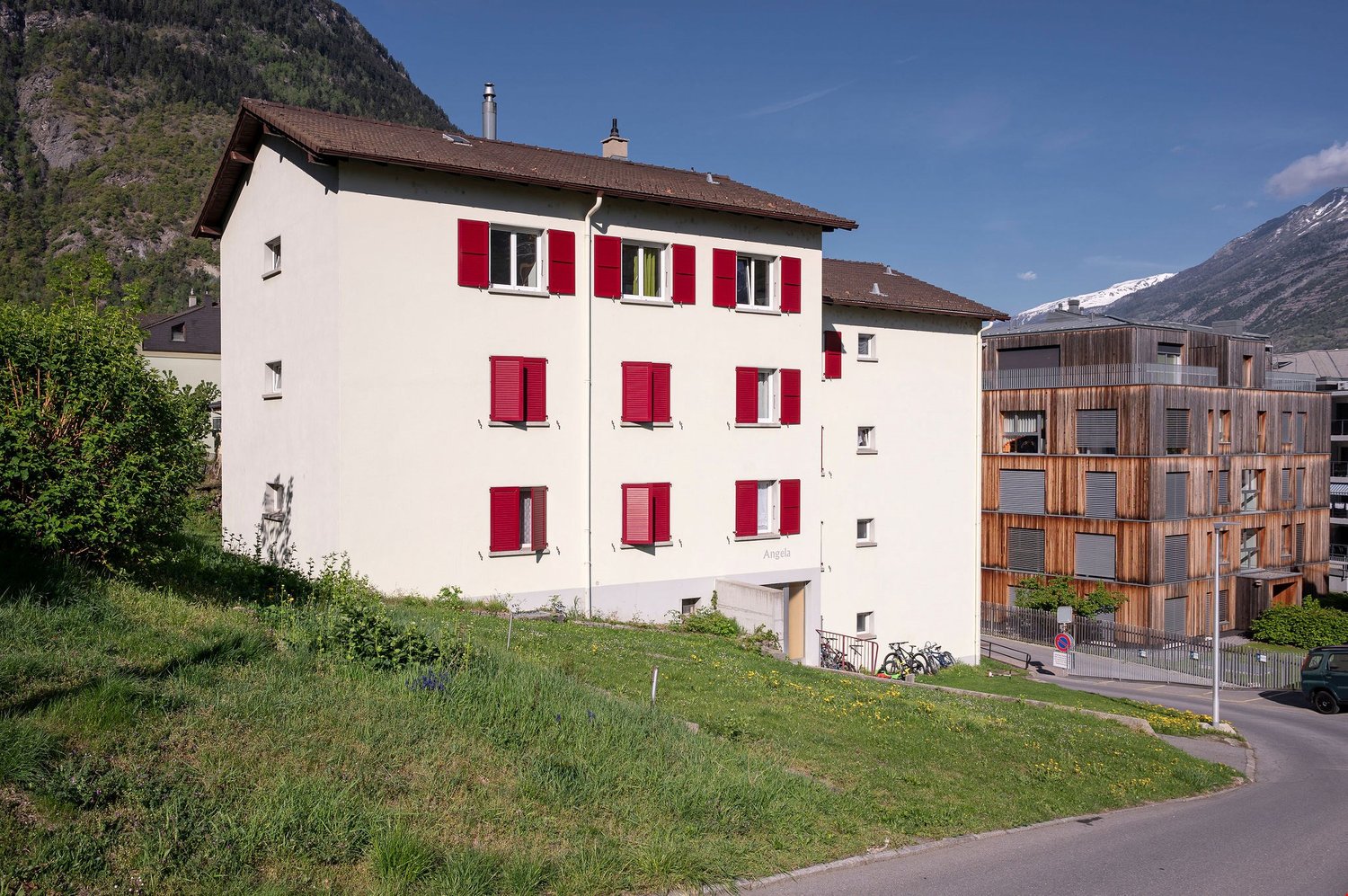 A multi-story residential building with a white exterior and red shutters. The building is situated on a grassy hill with a view of the surrounding mountains. There is a wooden building next to it.