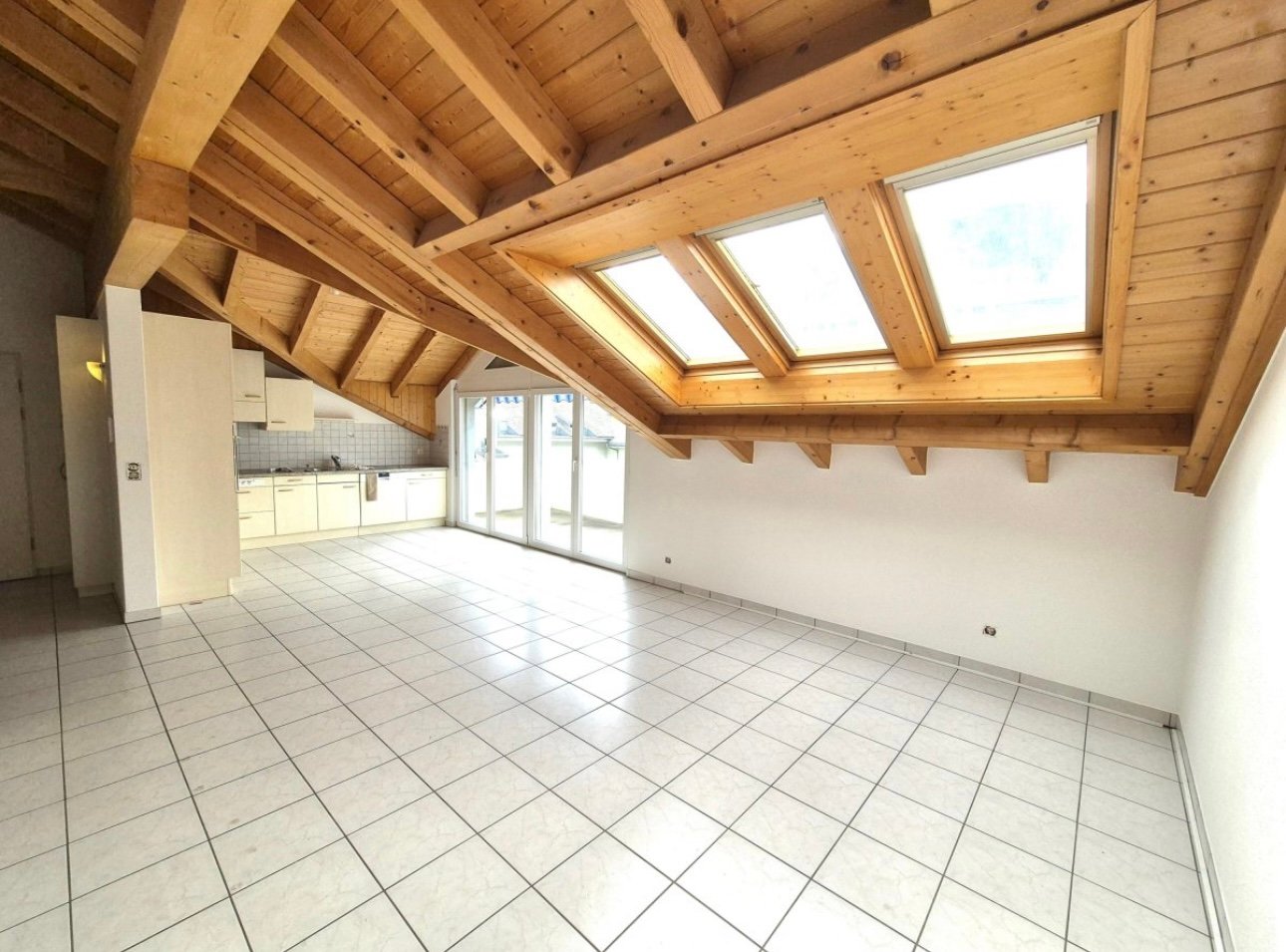 Spacious open-plan living area with vaulted wooden ceiling, large windows, and sliding glass doors leading to a balcony or terrace. The kitchen is located in the corner with white cabinets and tiled flooring throughout the space.
