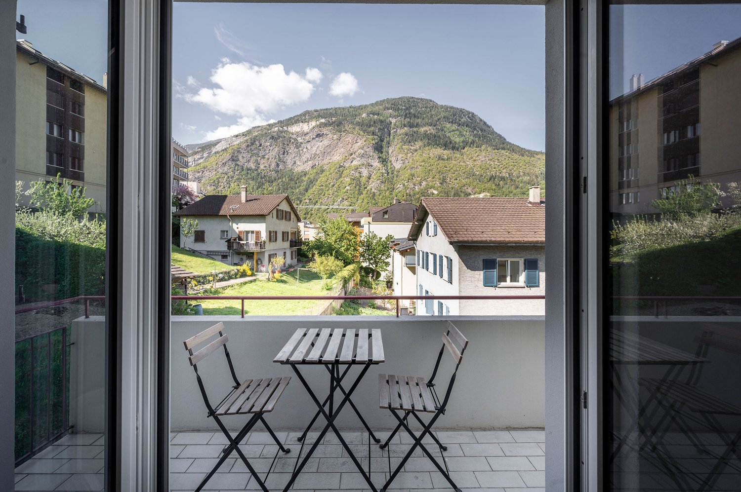 Balcony with a table and chairs overlooking a scenic mountain landscape, with residential buildings and greenery in the foreground