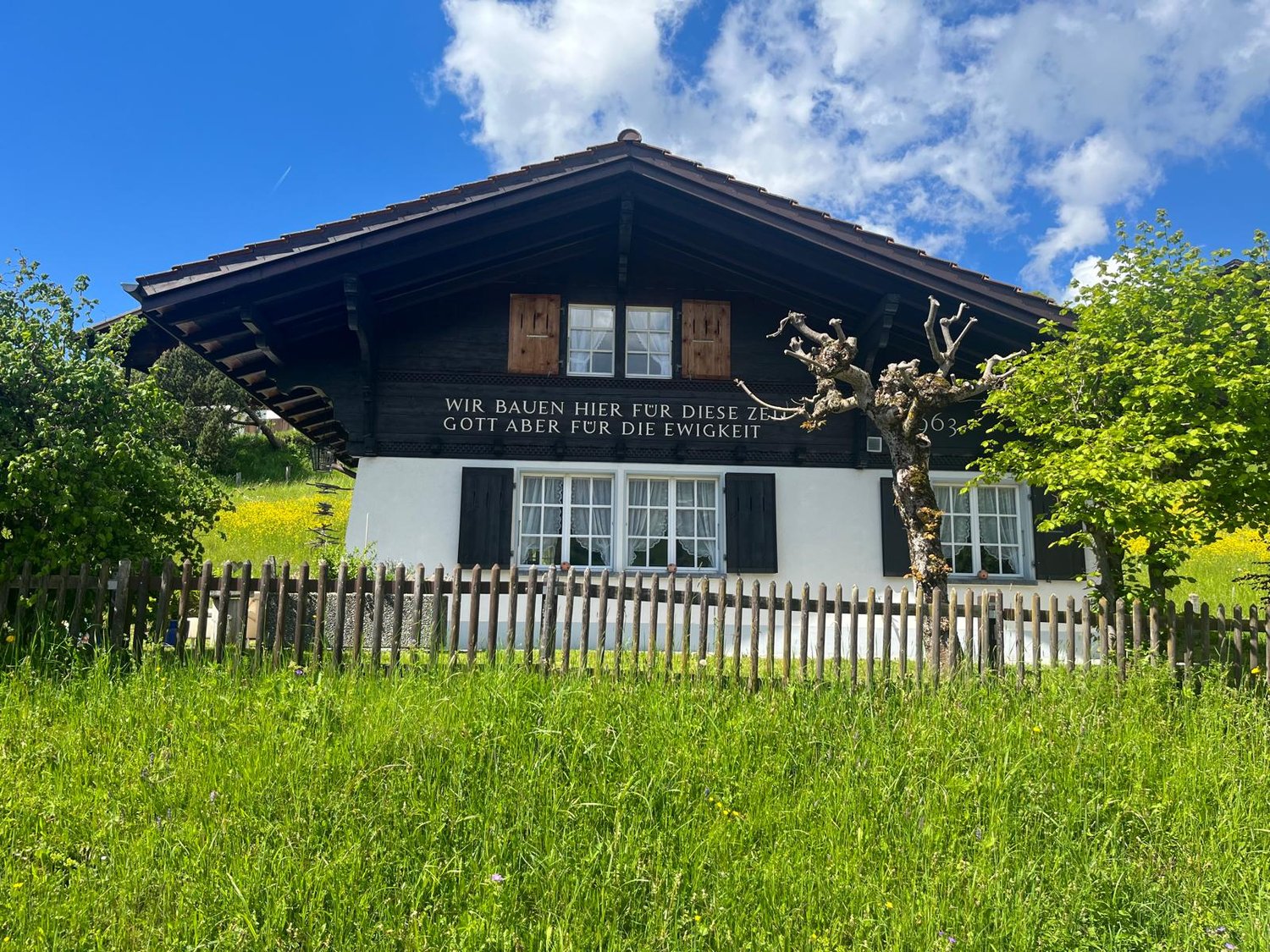 2 story chalet house with brown roof, white walls, wooden shutters, and wooden fence in front, surrounded by grass