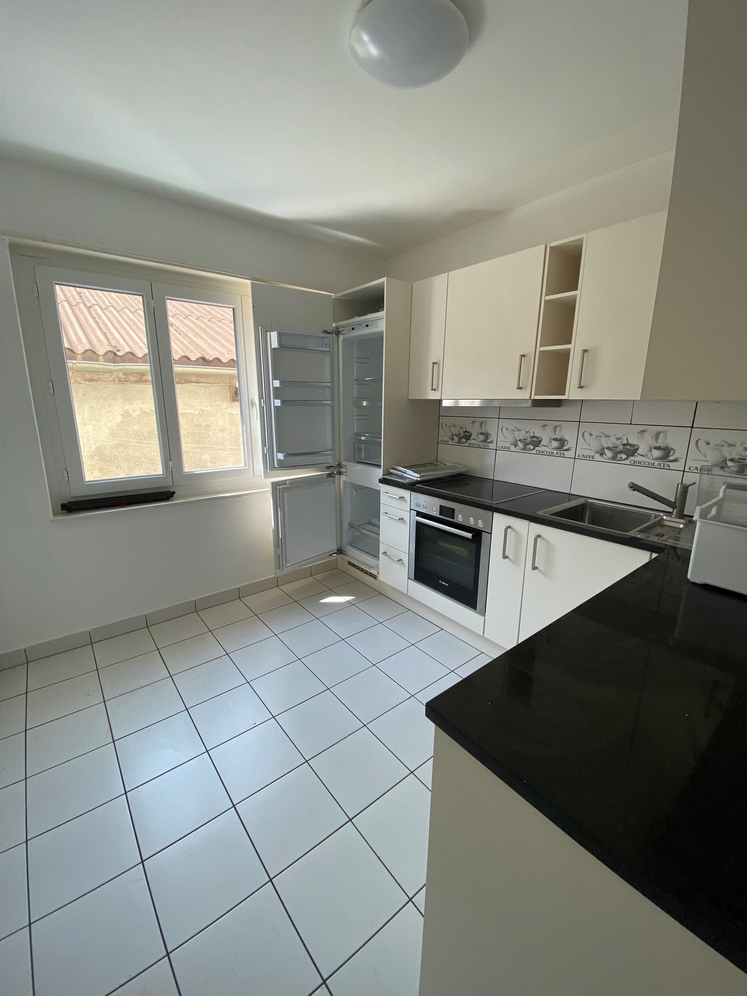 modern kitchen with white cabinets, tile floor, black countertop, built-in oven and refrigerator, sink with faucet, and a window
