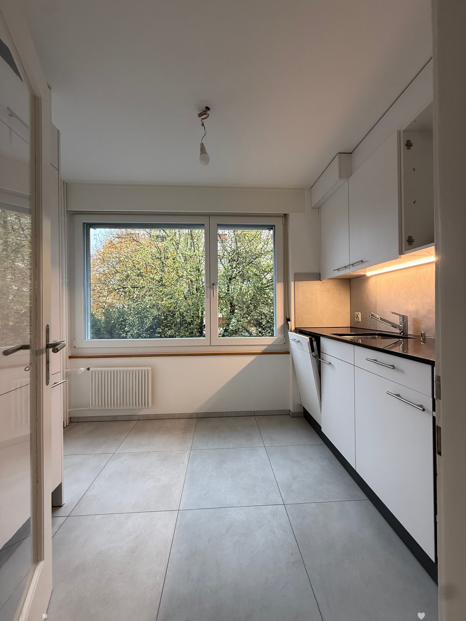 modern kitchen with large window, gray tiles, white cabinets, stainless steel sink, central light