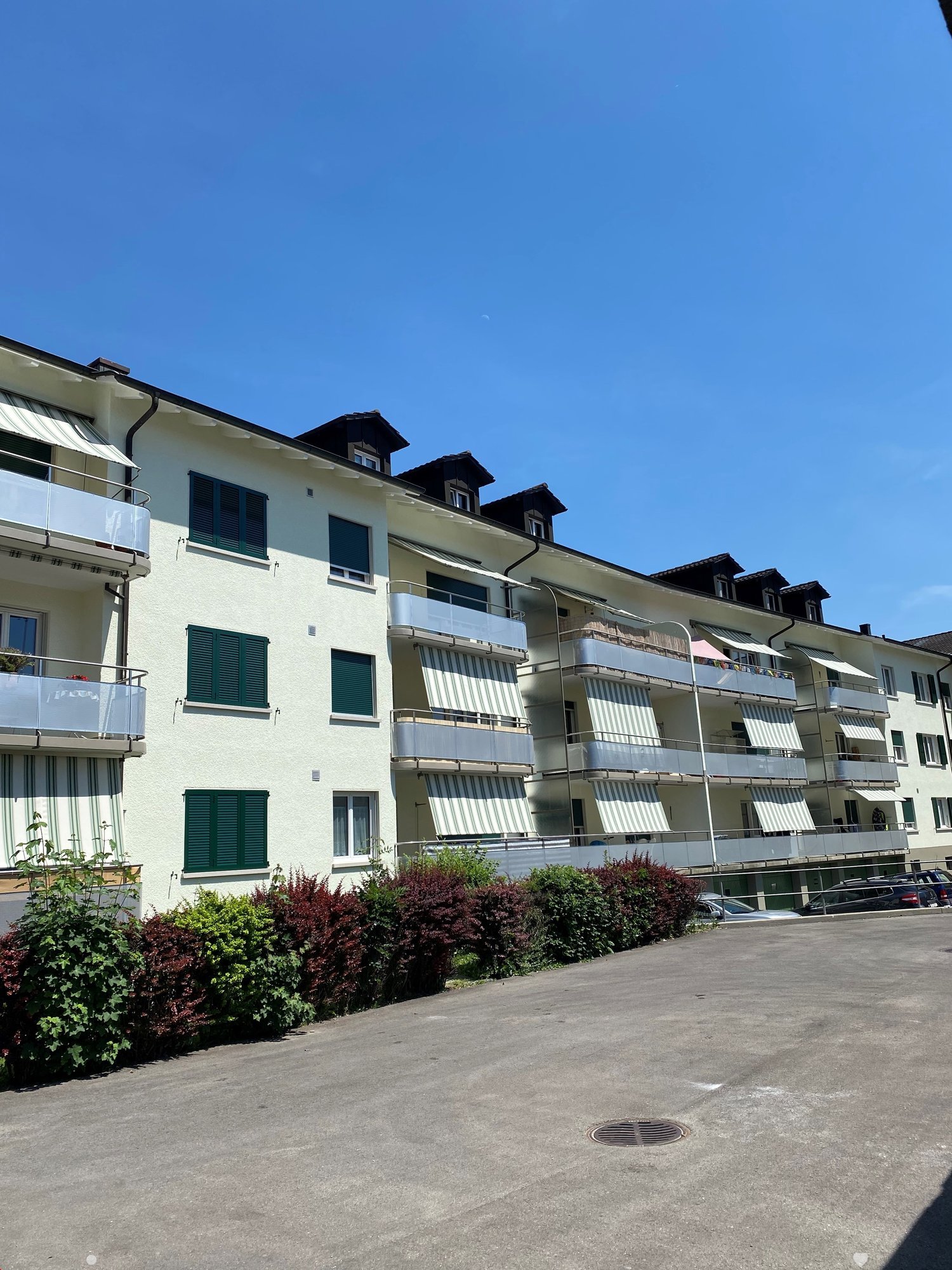 white apartment building with multiple balconies, several parked cars, greenery in front