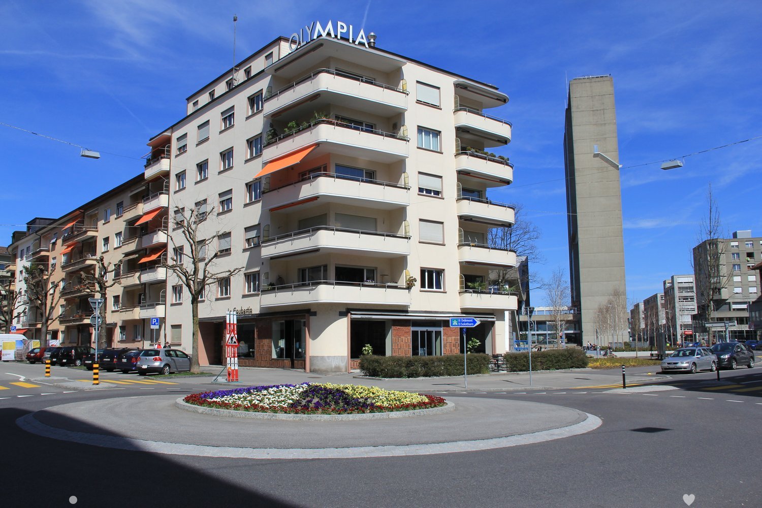 A multi-story building with the name 'OLYMPIAD' on top, surrounded by other buildings and trees.