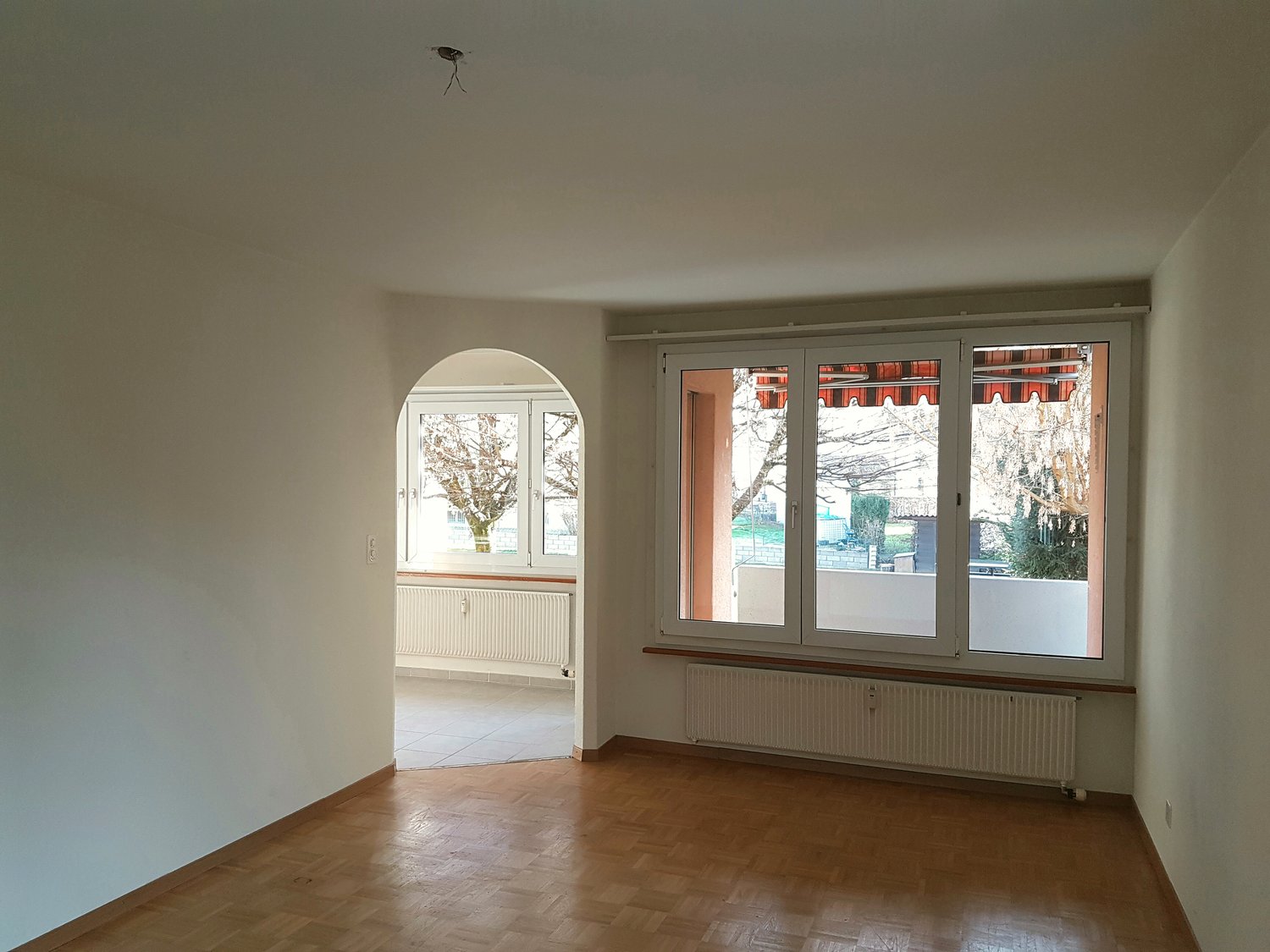 Spacious room with large windows, hardwood floors, and an arched doorway leading to another room. The room appears to be empty, with no furniture visible.