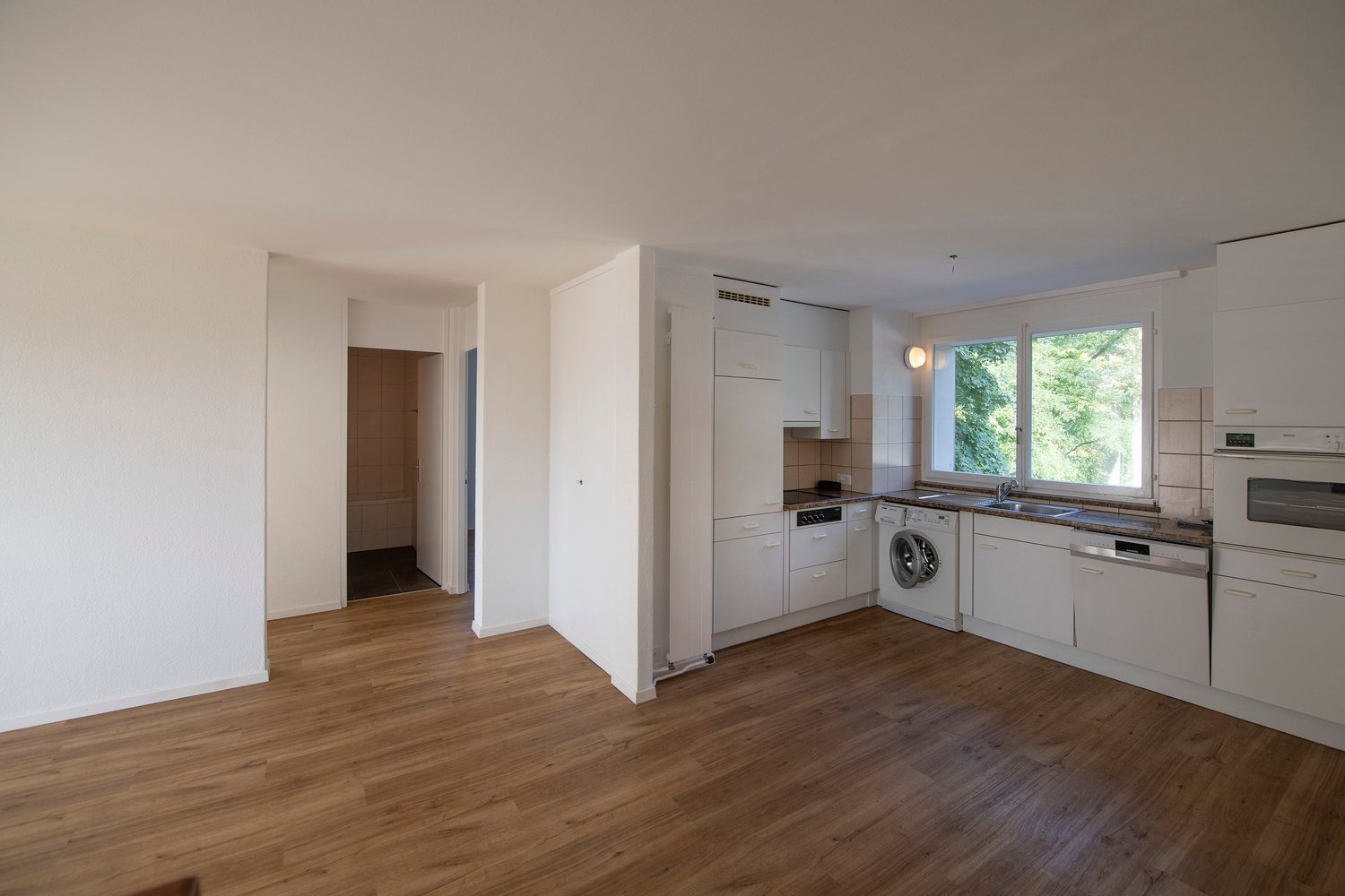 Open-concept living space with hardwood floors, white walls, and large windows overlooking greenery. The kitchen features modern white cabinets, appliances including a washing machine and dishwasher, and a sink. There is a hallway leading to other rooms.