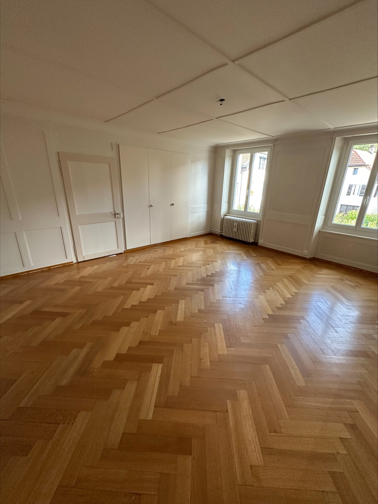 empty room, wooden parquet floor, white walls, wooden doors, radiator, two large windows