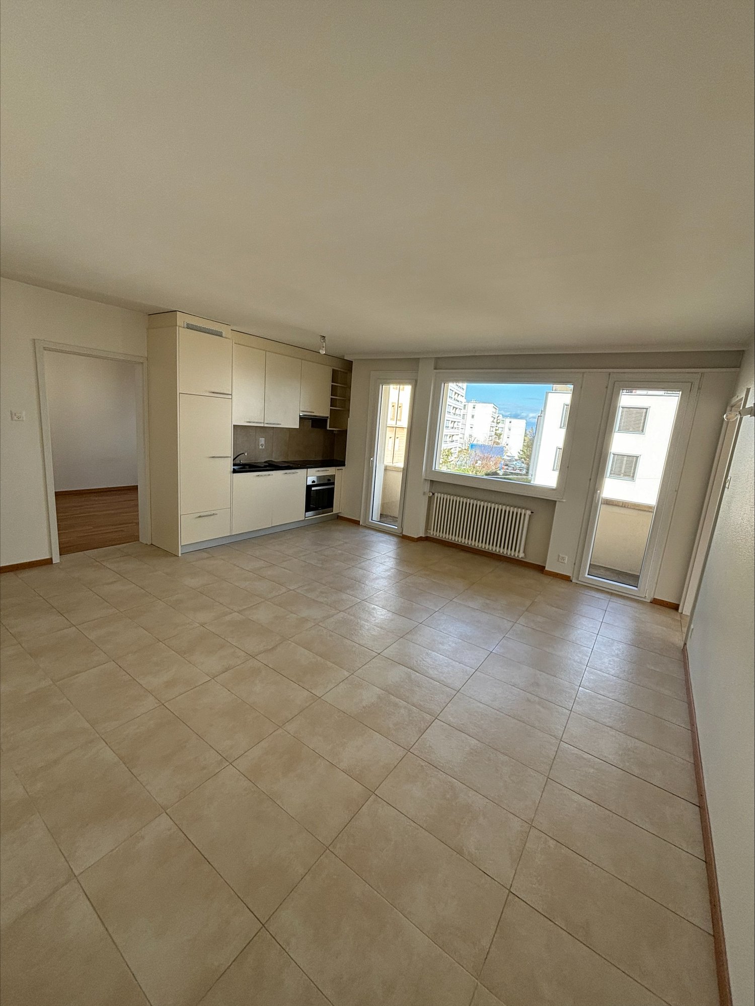 Empty room with tiled floor, white walls, kitchen area with sink, oven, and cabinets, sliding glass doors, and windows