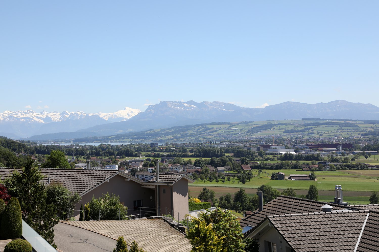 The image shows a scenic view of a town or city surrounded by mountains. The landscape includes residential buildings with red tile roofs, green fields, and a body of water in the distance. The mountains in the background appear snow-capped, indicating a