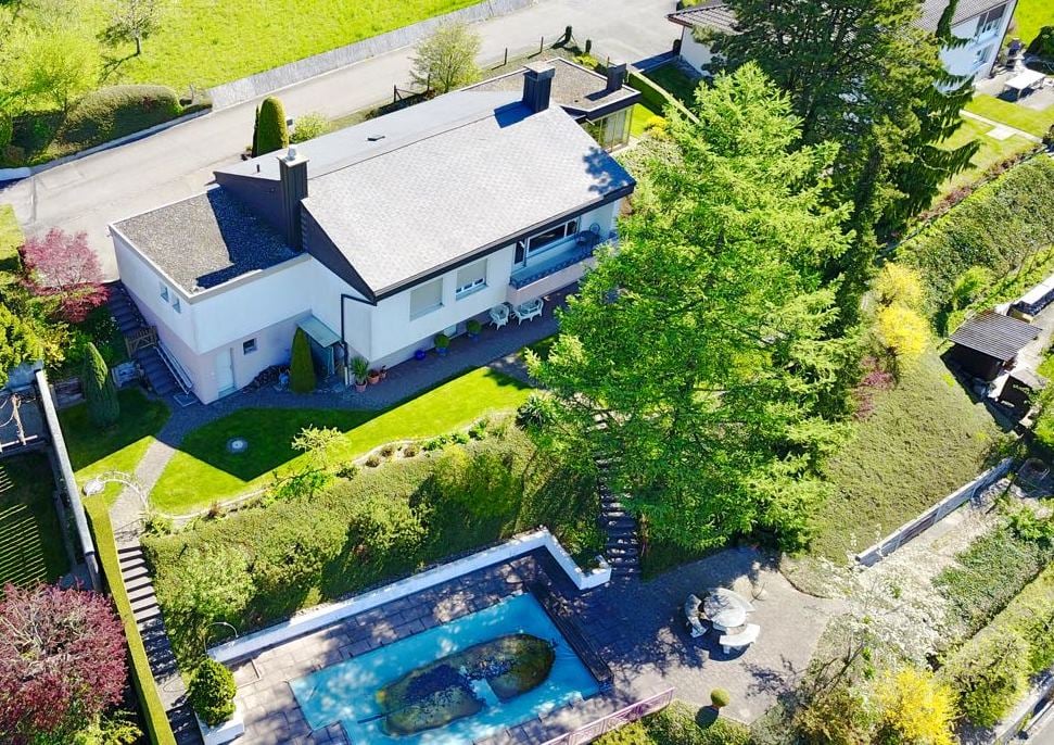 Aerial view of a 2-story house, gray roof, white walls, swimming pool, tree-lined garden, outdoor seating area, and road in the background