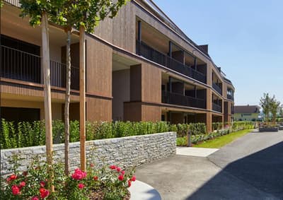 A modern apartment building with wooden facade, balconies, parking area, and greenery