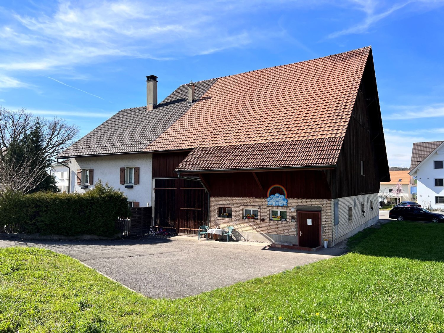 Red roof house, brick wall, wooden doors, large windows, plants, table and chairs, parked car, green lawn, and grass field.