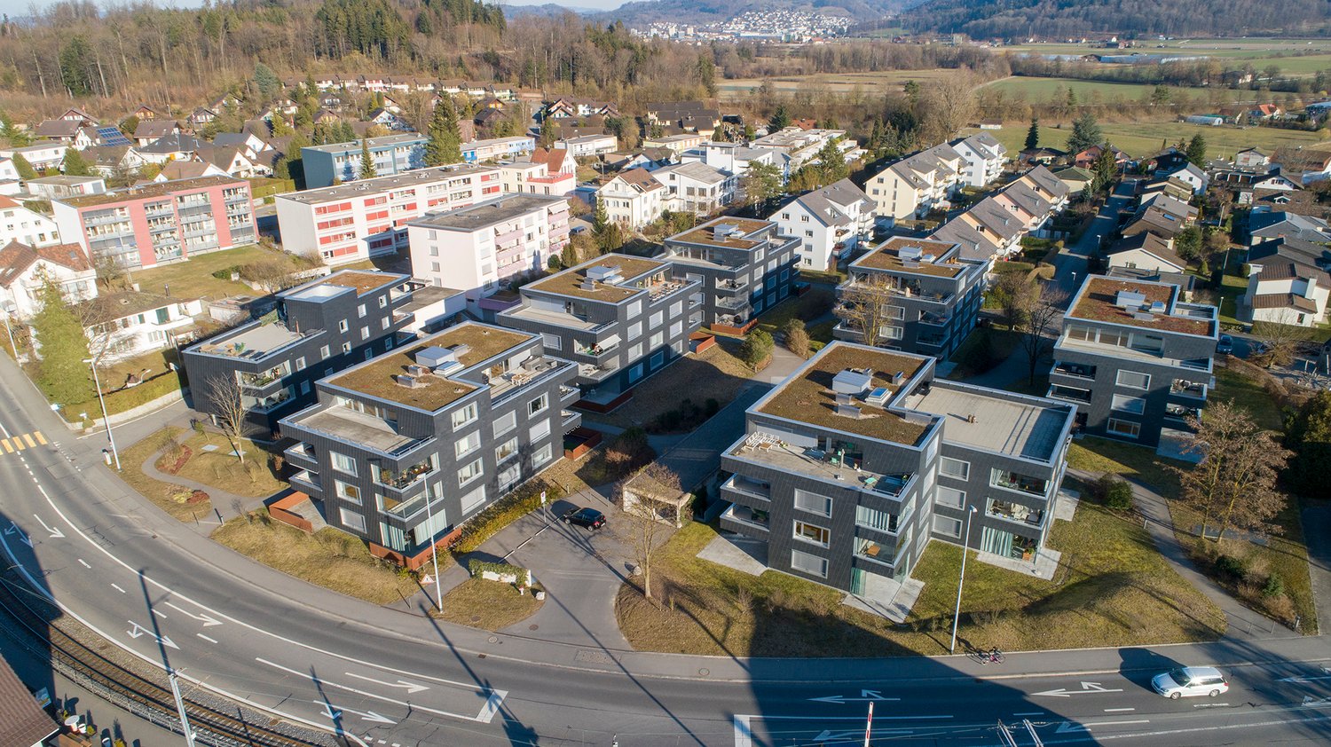 The image shows a residential area with multi-story apartment buildings, some with green roofs. The buildings are arranged in a compact layout, with a road running through the center. There appears to be a parking area visible in the foreground.