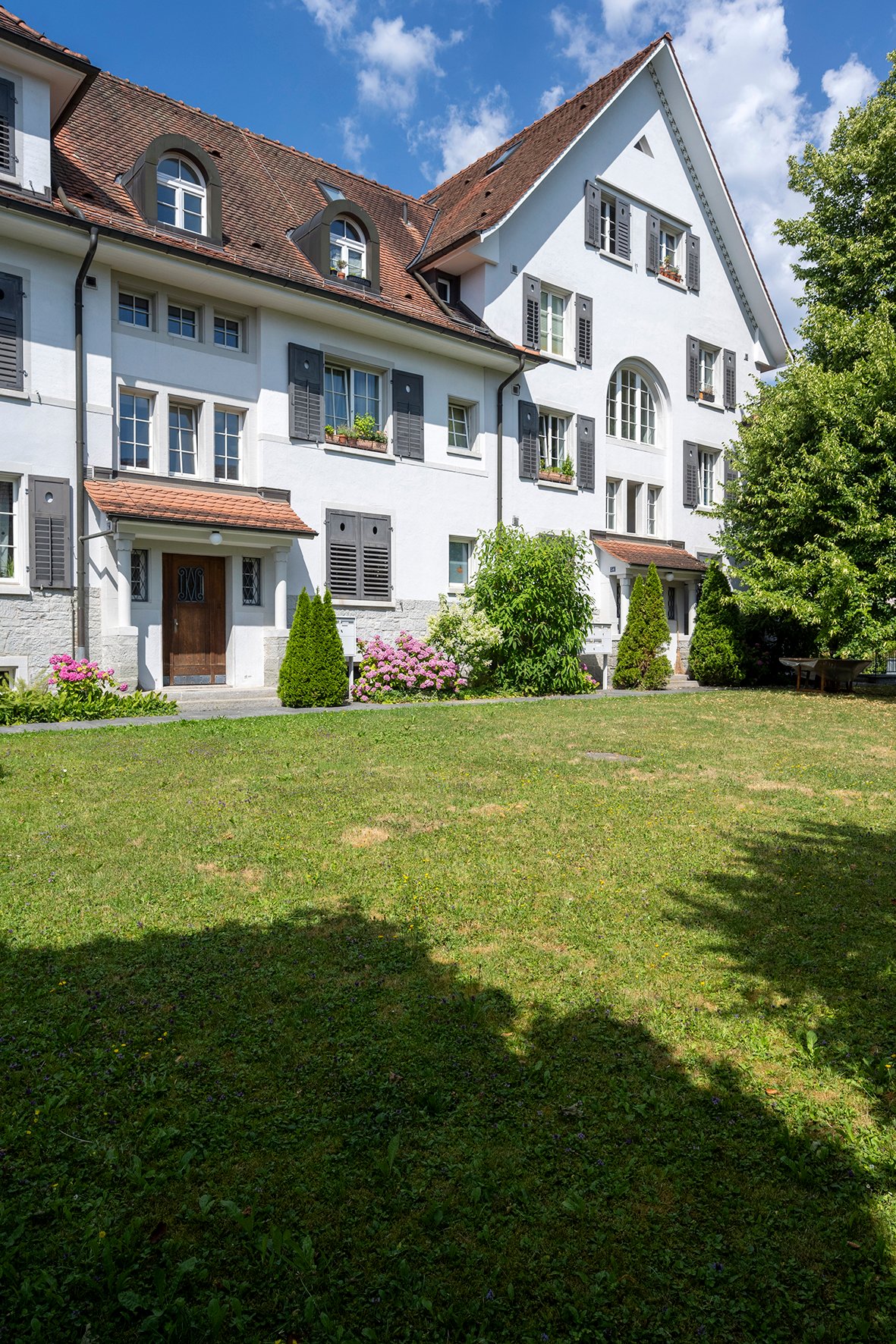 Exterior of a 2-story apartment building with red roof, white walls, multiple windows, brown wooden front door, and lush greenery in front
