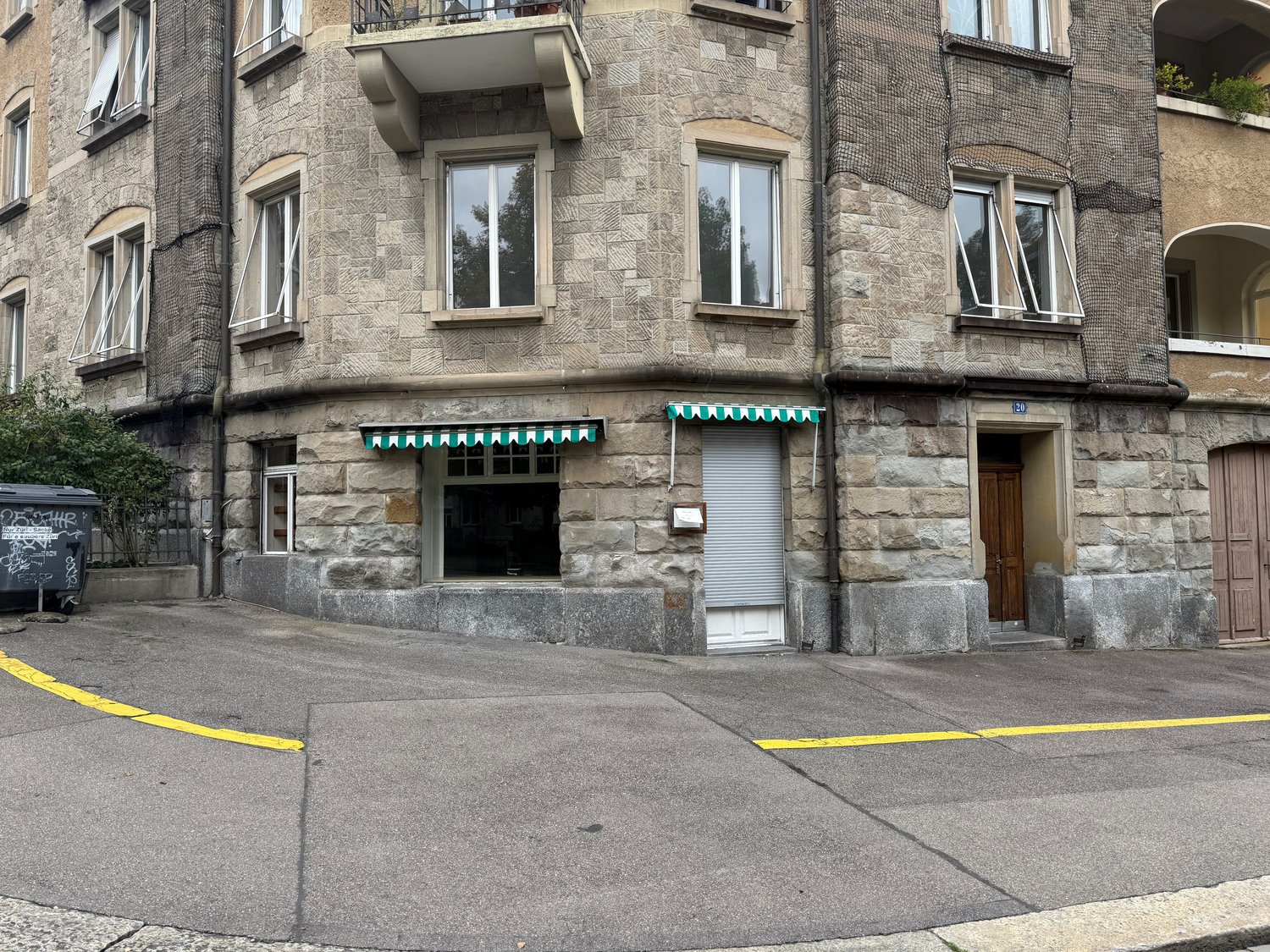 A stone building with a ground floor commercial space and residential apartments above. The building features multiple windows, a balcony on the second floor, and a roll-up door at the entrance of the commercial space.