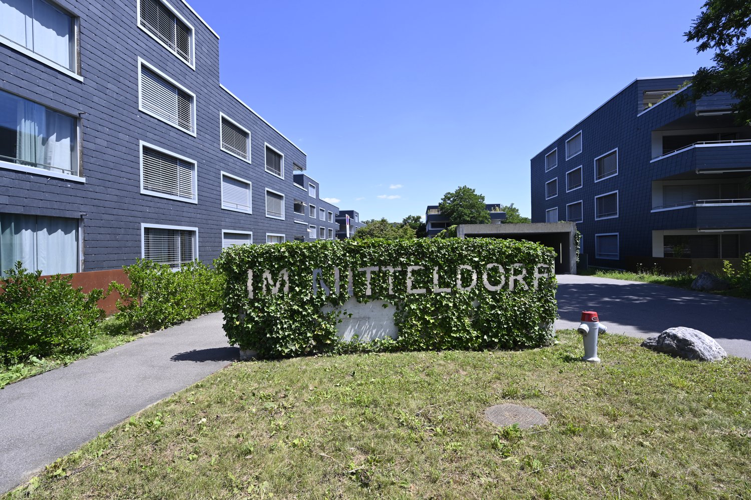 Entrance to a residential building complex with a sign reading 'IM MITTELDORF', surrounded by greenery and a pathway.