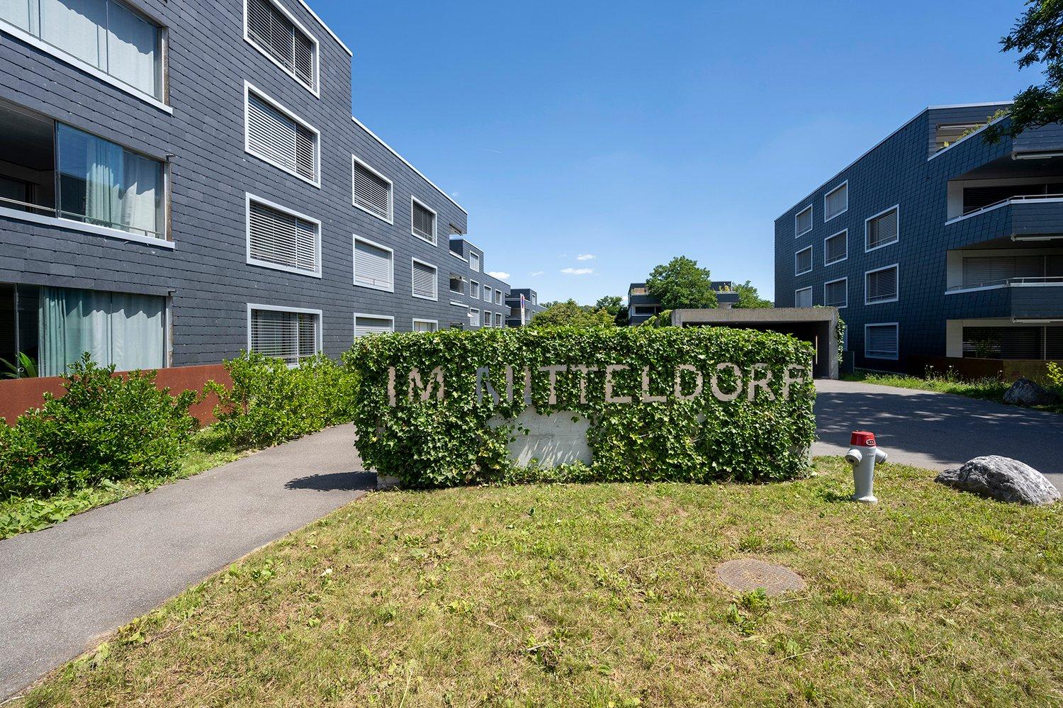 Modern apartment building with gray exterior, multiple windows, and a sign reading 'IM MITTELDORF'. The entrance is flanked by hedges and a fire hydrant. The pathway leads to the building with a clear blue sky in the background.