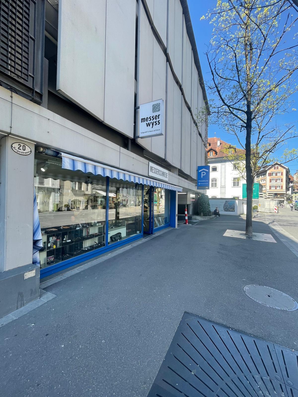 Commercial building with a shopfront, blue awning, and a sign reading 'Messer Wyss'. The building has a modern design with glass windows and a white wall. There is a parking sign and a tree in front of the building.