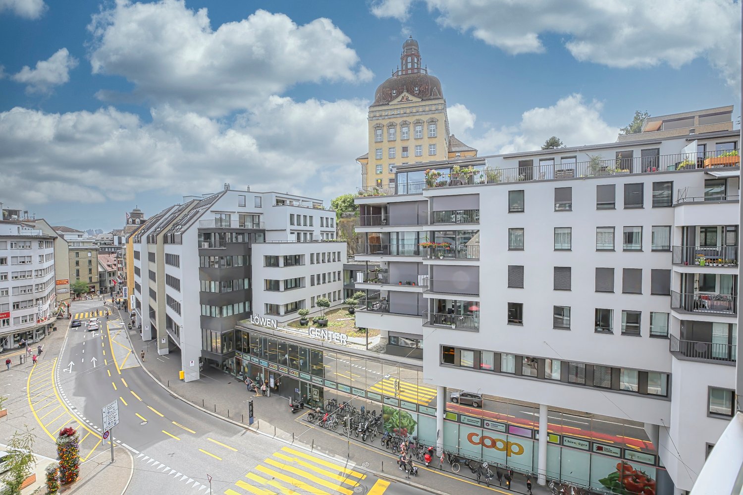 Aerial view of a modern building complex with multiple floors, glass facades, and balconies. The complex is situated in an urban setting with a busy street below, featuring bicycles parked along the sidewalk and a prominent building in the background.