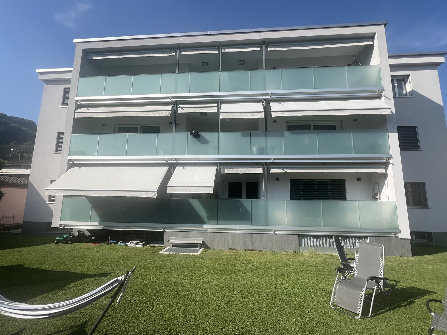 3-story modern apartment building with glass balconies, white exterior, and a grassy lawn in front