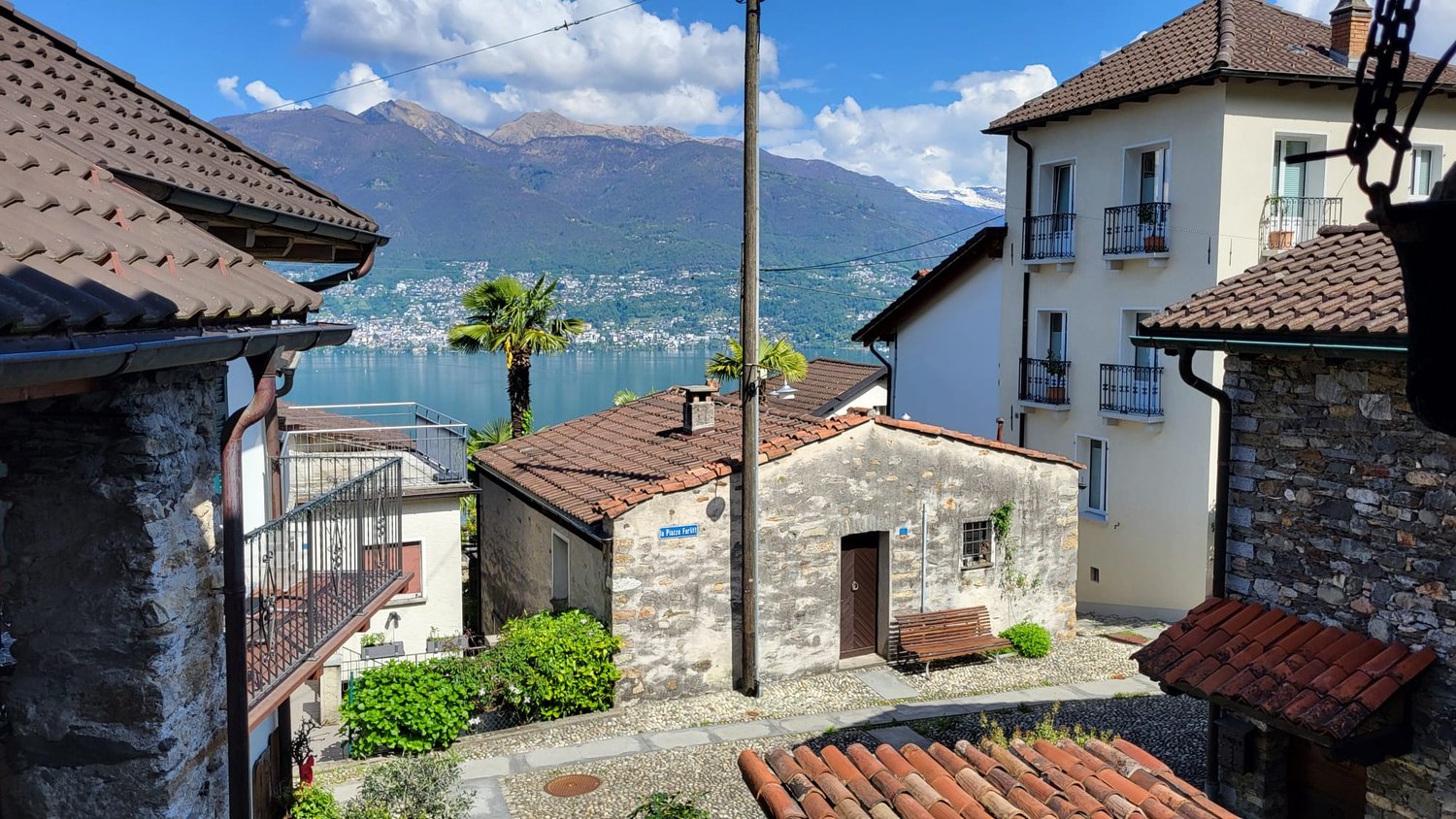 The image shows a picturesque view of a small town nestled along the shores of a lake, with mountains in the background. The buildings have traditional tile roofs and stone walls, and there are palm trees visible. The scene has a charming, quaint atmosphe