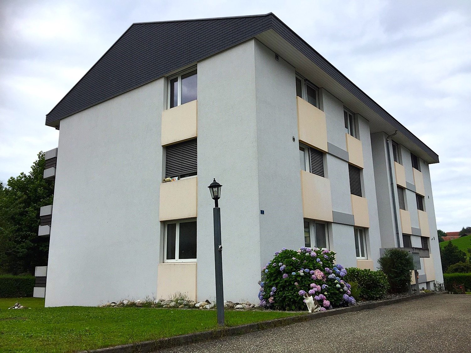 Apartment building, three stories, white and beige wall, grey roof, multiple windows, balcony with window on the second floor, street lamp, small garden in front