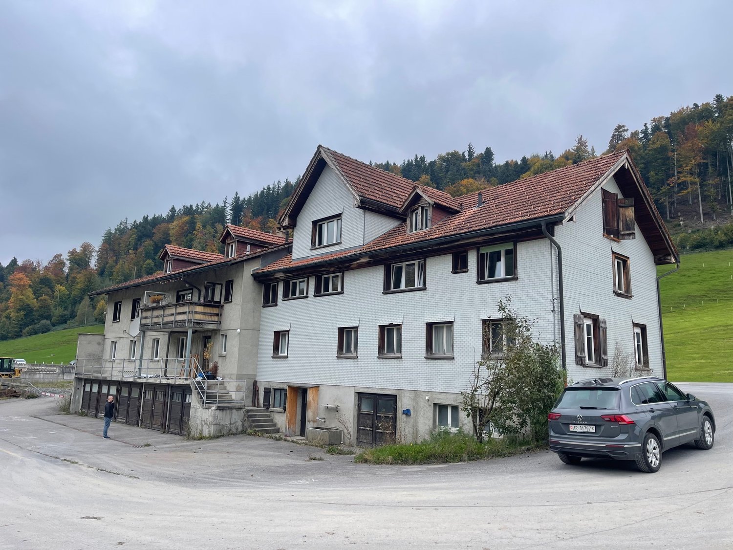 Large white house, brown roof, balcony, several windows, open door, parking area