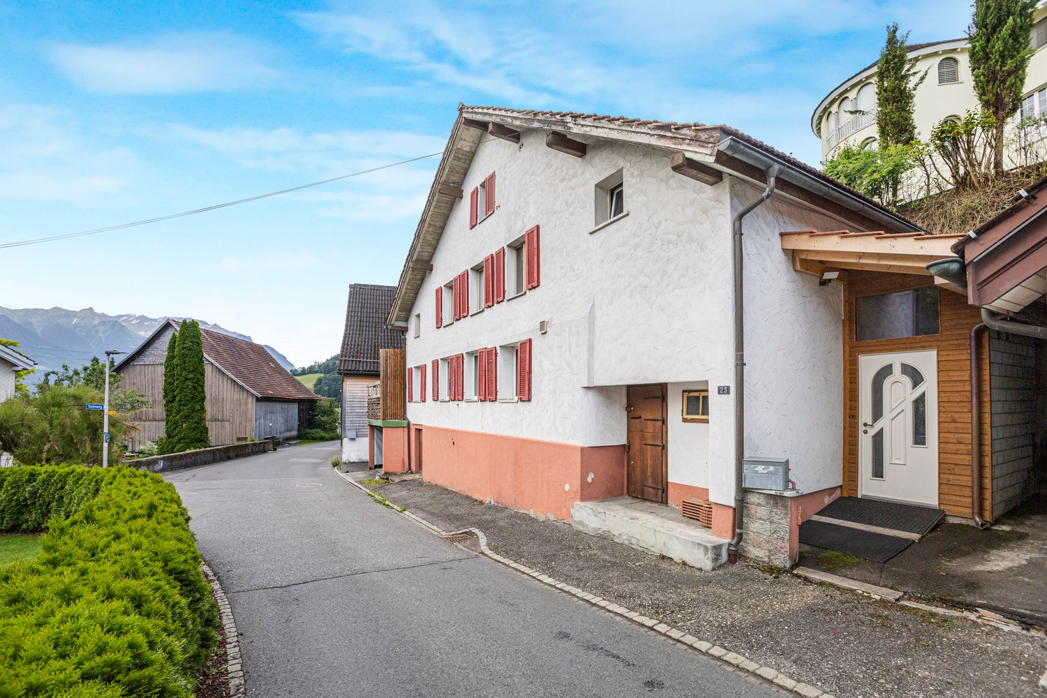 2-story residential building with white walls, red shutters, and a wooden roof. There is a gravel driveway leading to the building, and some trees and shrubs surrounding it. The building appears to be located in a rural or small town setting with mountain
