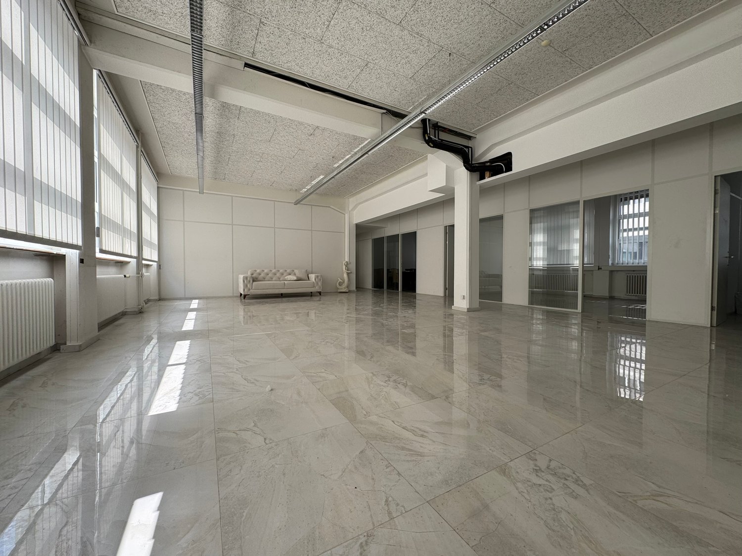 Large open office space with high ceilings, gray marble-like flooring, and a couch in the corner. The space appears to be empty and ready for use.