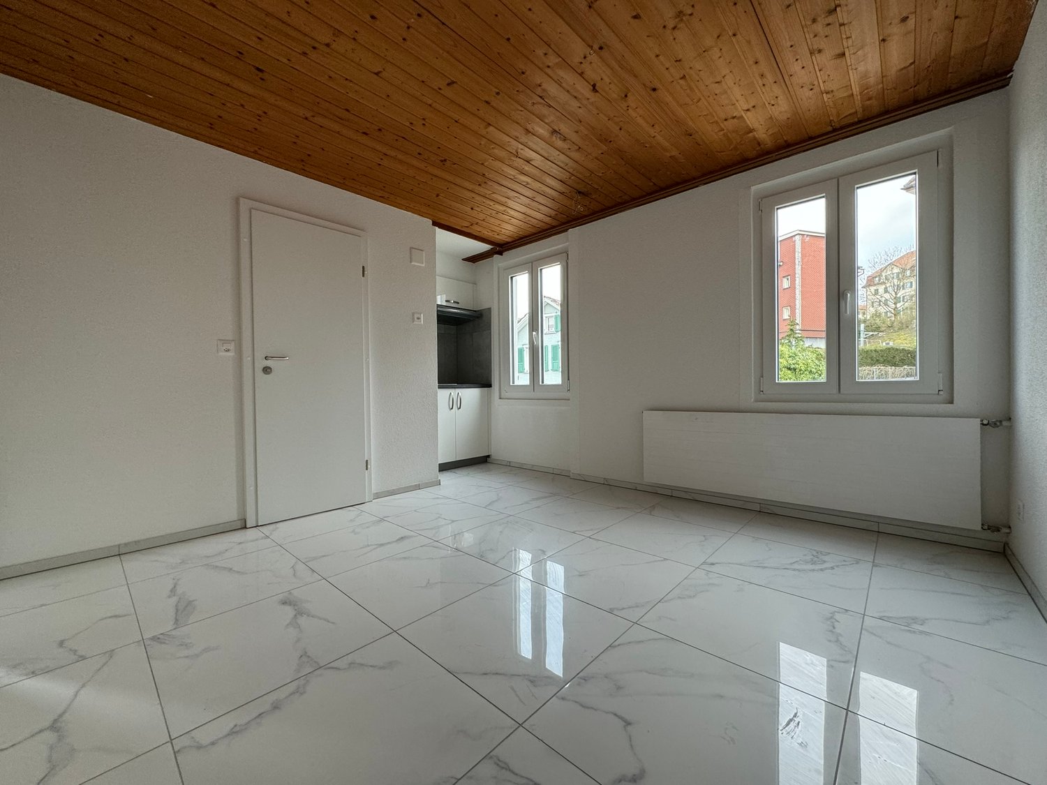 The image shows a studio apartment with a wooden ceiling, white walls, and large windows. The floor is covered with white marble-like tiles that reflect the light, creating a bright and spacious atmosphere. The room appears to be unfurnished, with only a