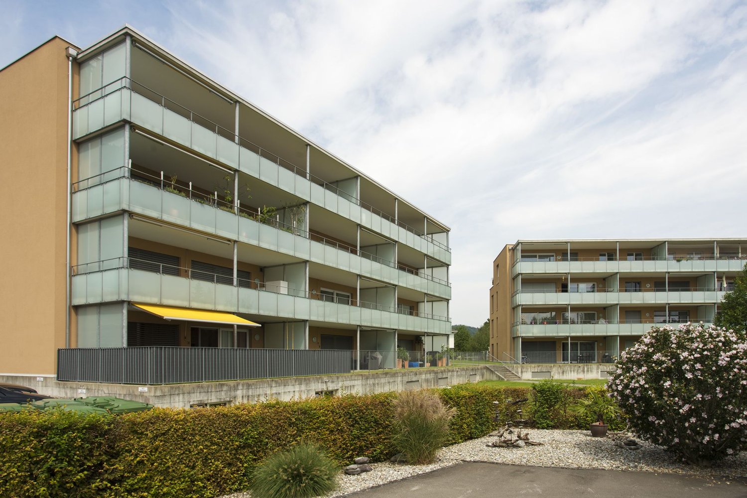 Apartment building, brown brick, green and white paint, balconies, yellow awning, garden with gravel and plants