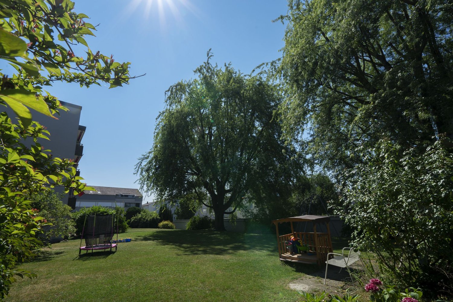Lush green lawn, mature trees providing shade, wooden benches and structures, residential buildings in the background