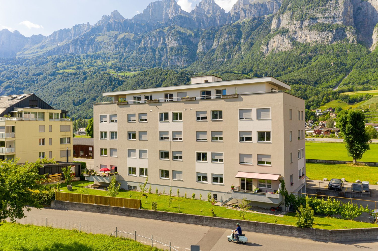 Multi-story apartment building with balconies, surrounded by green landscaping and with a view of the mountains in the background. A person riding a scooter is visible in the foreground.