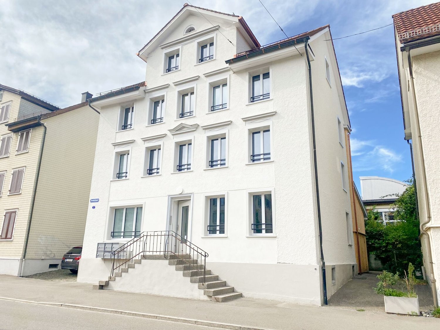 White building with multiple windows, stairs leading to the entrance, car parked in front, small garden with plants