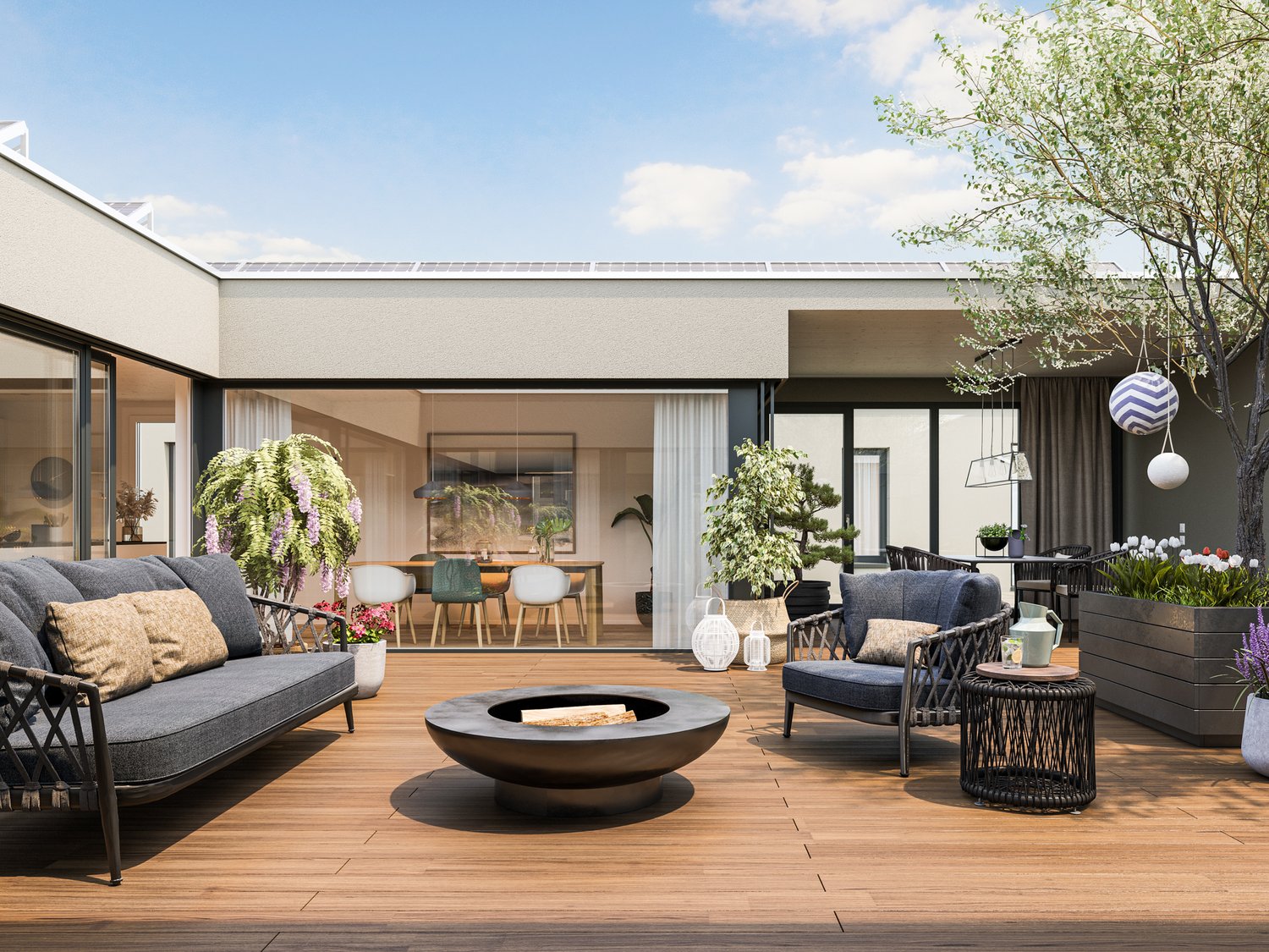 Spacious outdoor living area with a modern, minimalist design. The space features a large wooden deck, comfortable gray sofas and chairs, a circular fire pit, and potted plants and trees. The glass doors in the background suggest a connection to the indoo