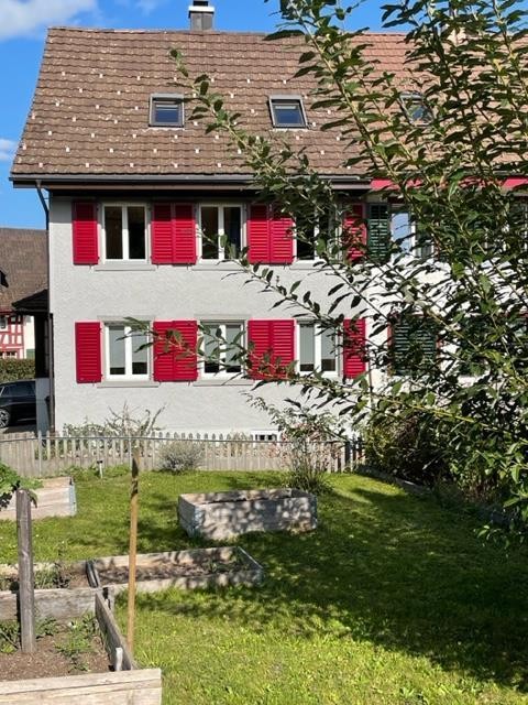 Two story house, red shutters, grey walls, brown roof, garden with wooden fences and plants